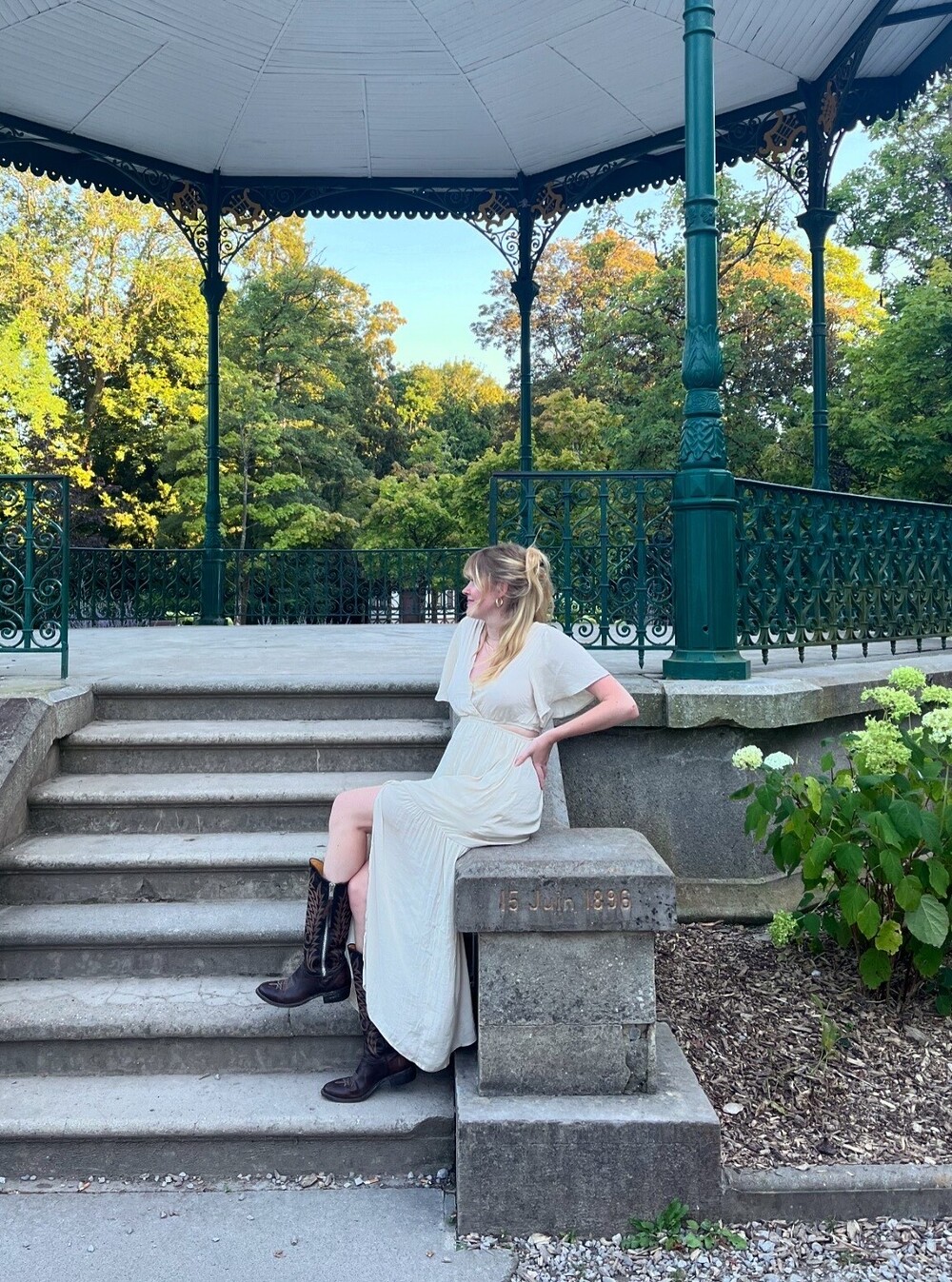 Oksana, gérante de Rembobine, sur des marches en robe blanche et bottes noires au jardin public de Saint-Omer