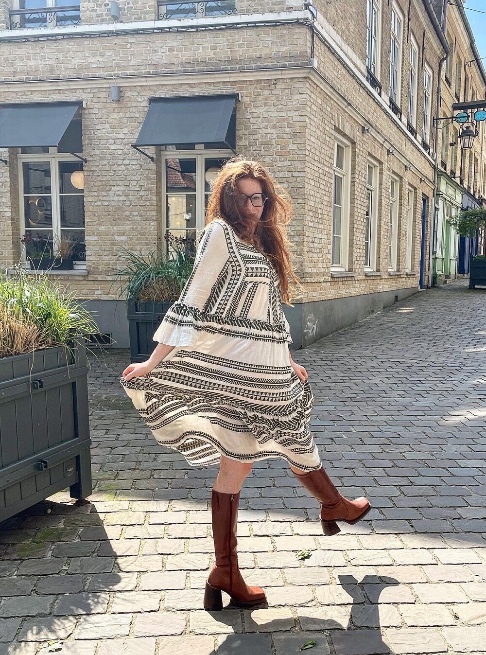 Une femme vêtue d'une robe et de bottes marrons, en plein soleil dans une rue