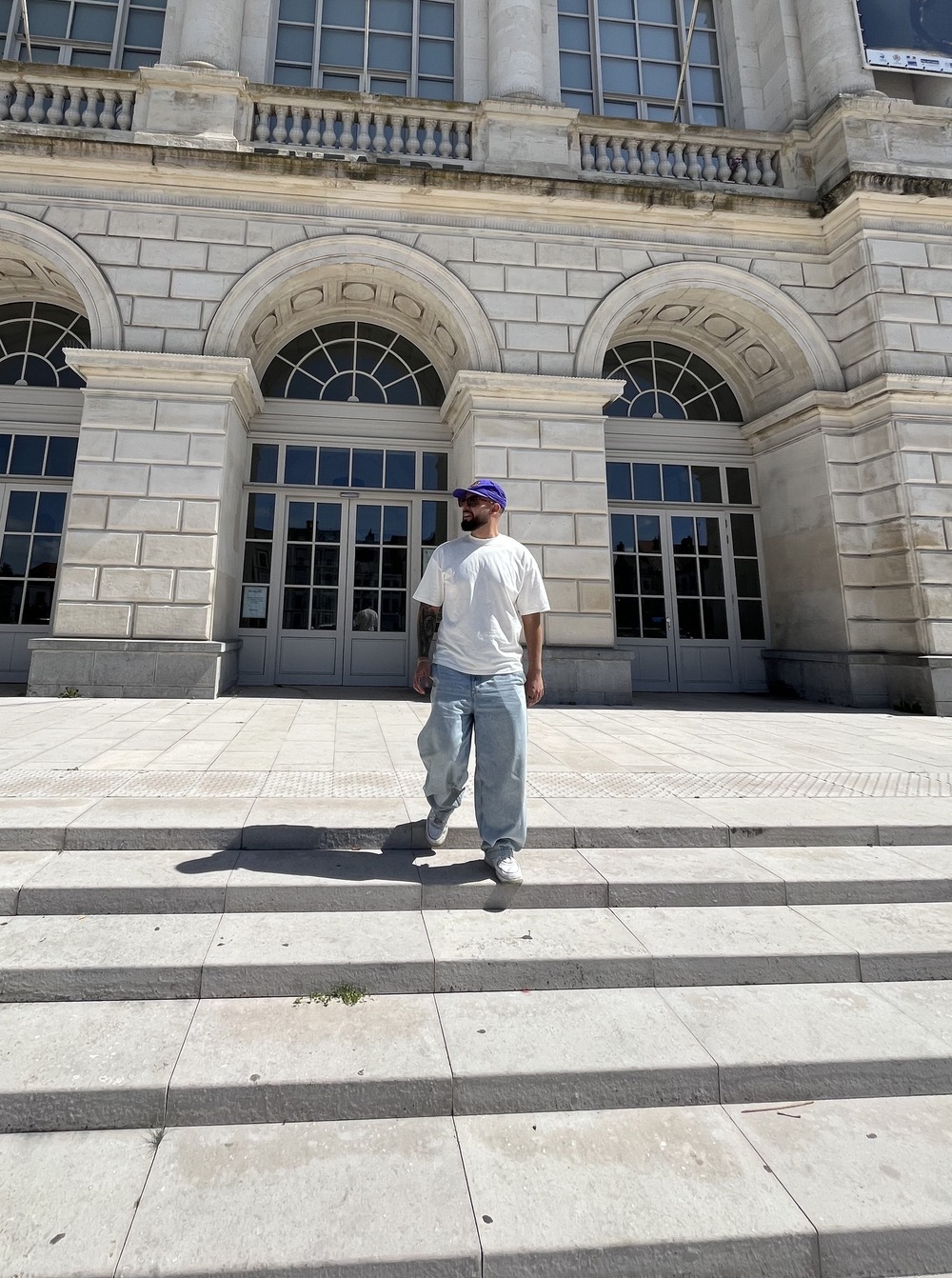 Un homme vêtue d'un t-shirt blanc et d'un jean large avec une casquette, sur des marches